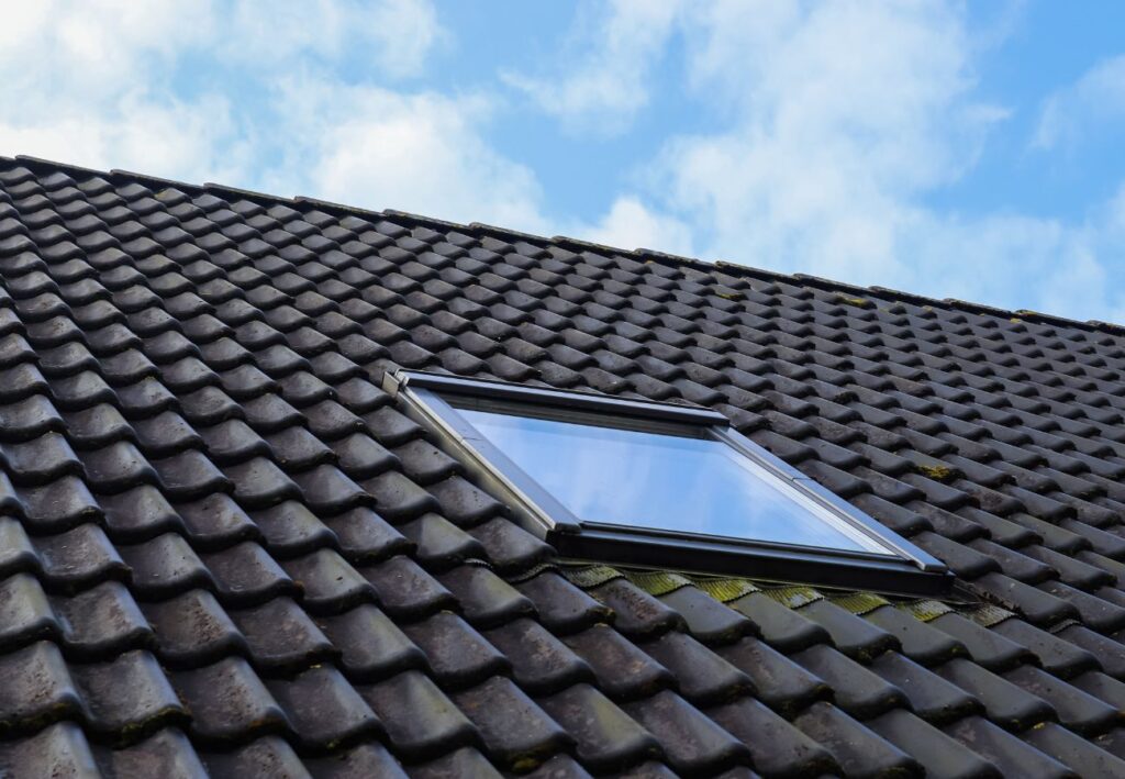 A skylight window installed on a sloped roof with dark tiles against a clear blue sky.